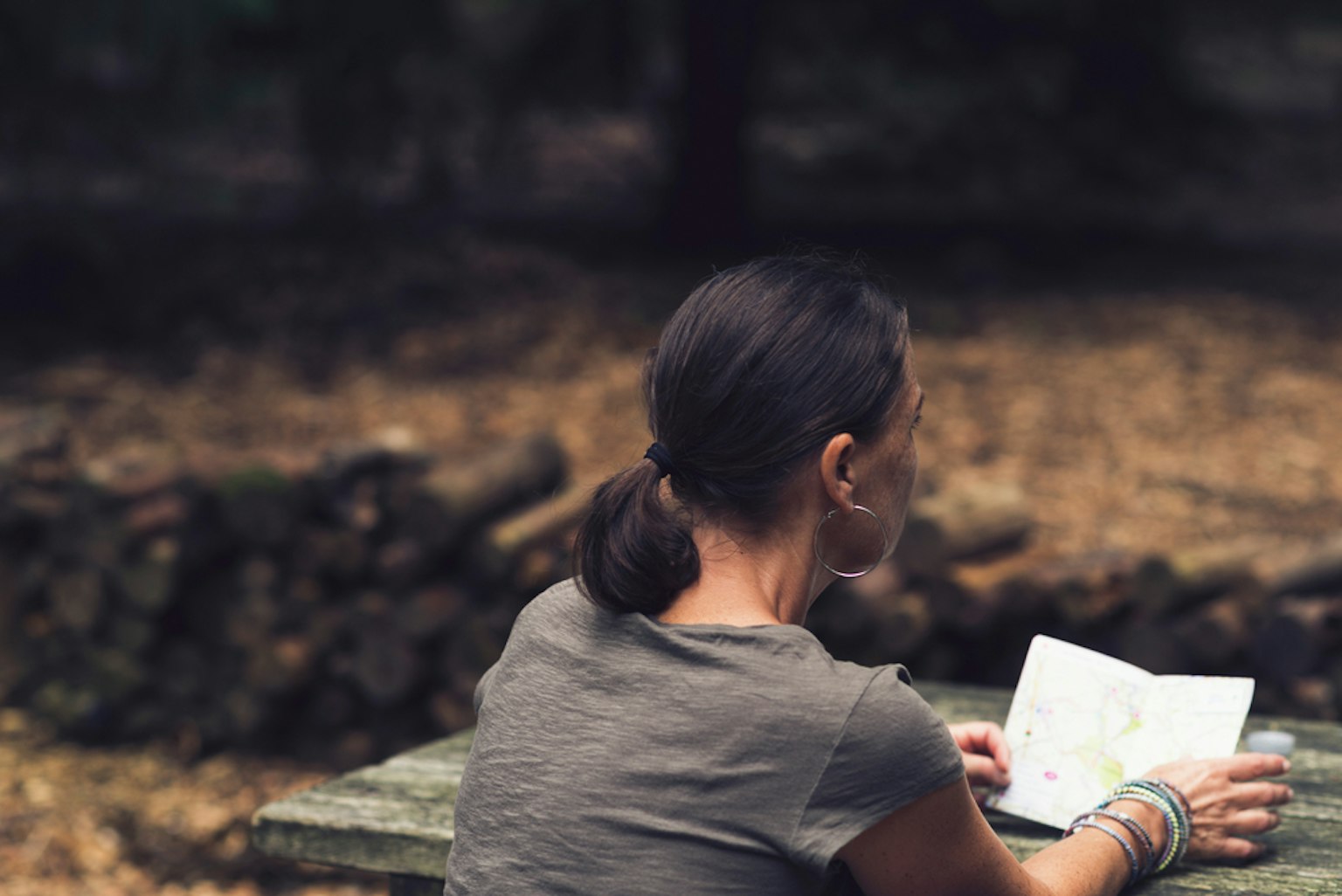 Rear View of Brunette Woman Reading Map at Wooden Table in Forest.